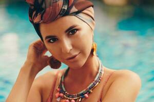 mulher com brilhante faço acima dentro boêmio roupa, turbante, étnico brincos, colar e pulseiras posando dentro natação piscina. foto