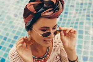 mulher com brilhante faço acima dentro boêmio roupa, turbante, étnico brincos, colar e pulseiras posando dentro natação piscina. foto