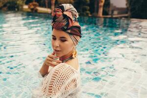 mulher com brilhante faço acima dentro boêmio roupa, turbante, étnico brincos, colar e pulseiras posando dentro natação piscina. foto