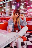 elegante menina dentro jeans Jaqueta sentado dentro t retro americano cafeteria. adorável sorridente mulher desfrutando doce leite mexe . vestindo branco tênis. foto