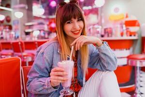 elegante menina dentro jeans Jaqueta sentado dentro t retro americano cafeteria. adorável sorridente mulher desfrutando doce leite mexe . vestindo branco tênis. foto