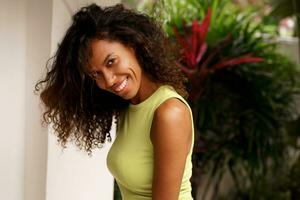 irradiando alegria e beleza, uma fechar-se retrato vitrines uma alegre africano americano mulher com Sombrio pele, ondulado cabelo, e uma cativante face. ela poses dentro uma tropical vila, abraçando a essência do verão Férias. foto