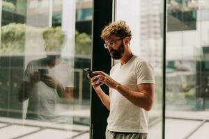 bonito homem com barba usando celular telefone enquanto caminhando dentro grande moderno cidade com arranha-céus. vestindo branco t camisa. foto