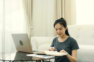 jovem mulher asiática senta-se na sala de estar usando uma calculadora para calcular a renda e as despesas da família e anota em seu notebook ou laptop em casa. foto