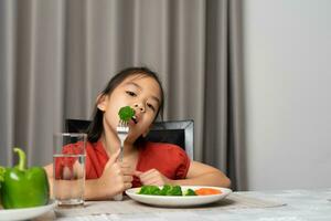 ásia pequeno menina comendo saudável legumes com saborear. foto