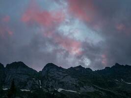 Sombrio silhuetas do montanhas contra uma brilhante Rosa pôr do sol céu. vermelho pôr do sol sobre majestoso montanhas. pôr do sol dentro magenta tons. atmosférico roxa panorama com uma grande altitude Nevado montanha vale. foto