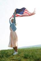 uma garota com um vestido coral e uma jaqueta jeans segura a bandeira dos EUA