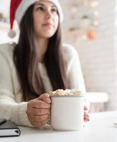 mulher morena com chapéu de Papai Noel segurando uma xícara de chocolate marshmallow