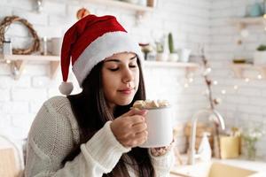 mulher com chapéu de Papai Noel segurando uma xícara de chocolate marshmallow na cozinha foto
