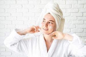 mulher caucasiana em toalha de banho branca aplicando creme facial em casa foto