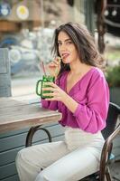 feliz morena menina com Rosa blusa sentado às a Barra , bebendo uma vidro do limonada enquanto sorridente às a Câmera. jovem sexy bonita mulher em a cadeira bebendo suco vestindo uma branco calça foto