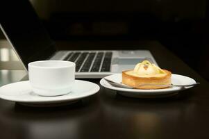 ainda vida com Comida e eletrônico dispositivo. uma doce delicioso tortinha, copo do café sobre computador portátil fundo. foto