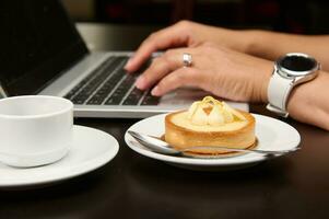 fechar-se delicioso tartelete com limão creme sobre a fundo do fêmea mãos digitando texto em computador portátil teclado. foto