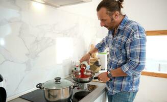 atraente masculino chefe de cozinha mão adicionando passata para dentro uma fritar panela com Oliva óleo e baía folha, cozinhando italiano massa para jantar foto