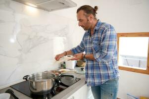 bonito jovem homem colocando picado alho a partir de corte borda para dentro uma panela com ebulição tomate molho, cozinhando às casa foto