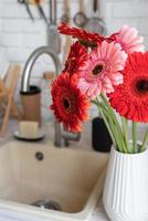 margaridas gerbera vermelha e rosa em um vaso branco em uma cozinha de madeira foto