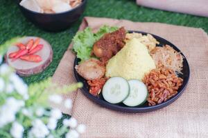 indonésio estilo amarelo arroz com frango e lado pratos dentro Preto prato em verde Relva fundo foto