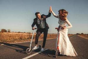 casal de noivos sorridente andando de scooters ao longo da estrada ao pôr do sol foto