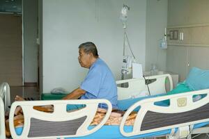 a idosos ásia masculino paciente senta em uma hospital cama esperando para uma médico para Verifica em ele durante dele hospital ficar. suave e seletivo foco. foto