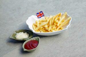 francês fritas dentro uma prato decorado com Reino Unido bandeira. Está batata cunhas frito dentro óleo, crocantes em a lado de fora e suave em a dentro. borrifar com sal e servir enquanto quente.comer com ketchup e maionese foto