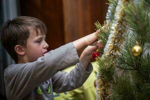 Garoto se apega uma vermelho bola em uma ramo do uma Natal árvore às lar. preparando para Natal às lar. real pessoas. decoração Natal árvore com uma variedade do brinquedos foto