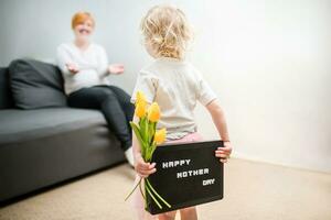pequeno menina detém uma ramalhete do amarelo tulipas e uma placa este diz feliz mãe dia. criança dá flores para mãe para feriado. foto