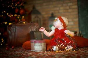 pequeno menina dentro vermelho vestir contra fundo do Natal árvore detém Natal festão dentro dela mãos. bebê 6 mês velho comemora Natal. foto