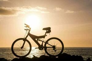 uma bicicleta é em pé em uma Rocha perto a oceano às pôr do sol foto
