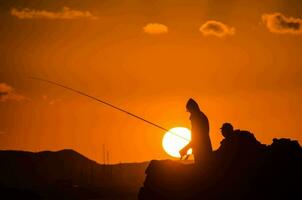 dois pessoas pescaria às pôr do sol em uma Rocha foto