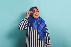 animado meia idade ásia mulher dentro azul hijab e listrado camisa mostrando, apresentando crédito cartão para pagando conectados negócios, isolado sobre azul fundo. foto