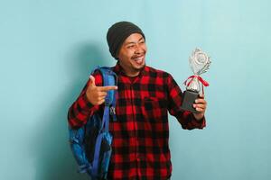 animado jovem ásia homem aluna orgulhosamente apontando uma troféu dentro dele mão, isolado em azul fundo foto