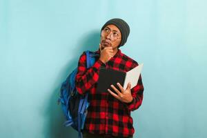 pensativo jovem ásia aluna homem é pensando enquanto segurando livro e em pé contra azul fundo foto