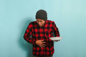 descontente jovem ásia homem com uma gorro chapéu e uma vermelho xadrez flanela camisa é dando a Comida uma mau análise, enquanto segurando a esvaziar branco placa, isolado em uma azul fundo foto