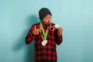 animado jovem ásia homem com uma gorro chapéu e uma vermelho xadrez flanela camisa é espantado, com dele boca aberto dentro surpresa e descrença, Como ele vitórias medalhas enquanto em pé contra uma azul fundo foto