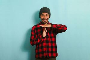 jovem ásia homem com gorro chapéu e vermelho xadrez flanela camisa sorridente às a Câmera enquanto fazer uma tempo esgotado gesto, isolado em uma azul fundo foto