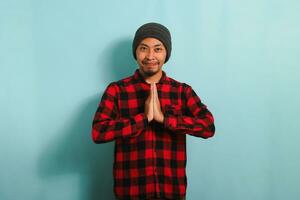 uma jovem ásia homem com uma gorro chapéu e uma vermelho xadrez flanela camisa gestos a eid Mubarak cumprimento e congratula-se com Ramadã enquanto em pé contra uma azul fundo foto