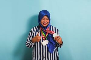 a animado meia idade ásia mulher dentro uma azul hijab e uma listrado camisa é dando uma afirmativo enquanto segurando uma medalha, isolado em uma azul fundo foto