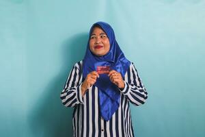 animado meia idade ásia mulher dentro azul hijab e listrado camisa mostrando, apresentando crédito cartão para pagando conectados negócios, isolado sobre azul fundo. foto