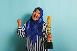 uma orgulhoso meia idade ásia empresária dentro uma azul hijab e uma listrado camisa é fazer uma vencedora gesto enquanto segurando uma ouro troféu, a comemorar dela sucesso. ela é isolado em uma azul fundo. foto