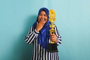 a animado meia idade ásia empresária dentro uma azul hijab e uma listrado camisa é segurando uma ouro troféu, a comemorar dela sucesso e conquista. ela é isolado em uma azul fundo foto