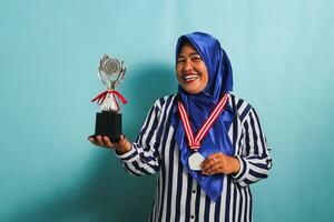 uma feliz meia idade ásia empresária dentro uma azul hijab e uma listrado camisa é mostrando a esvaziar branco medalha enquanto segurando uma prata troféu, a comemorar dela sucesso, isolado em uma azul fundo foto