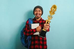 feliz jovem ásia homem vestindo uma mochila, gorro chapéu, e vermelho xadrez flanela camisa, segurando uma ouro troféu, alegria dentro dele sucesso e conquista, isolado em uma azul fundo. foto