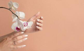 mãos do uma jovem mulher com branco grandes unhas em uma bege fundo com orquídea flores manicure foto