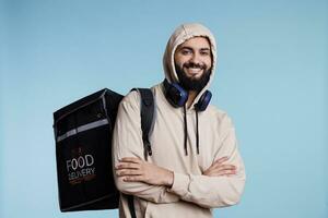 árabe homem com alegre expressão carregando mochila e entregando Comida retrato. jovem sorridente restaurante entregador vestindo de capuz em pé com térmico saco e olhando às Câmera foto