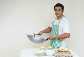 bonito ásia homem chefe de cozinha é cozinhando frito ovos, desgasta branco camisa e avental, detém fritar panela e concha espátula . conceito, amor culinária. cozinha estilo de vida. foto