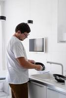 homem de camiseta branca lavando pratos na cozinha foto