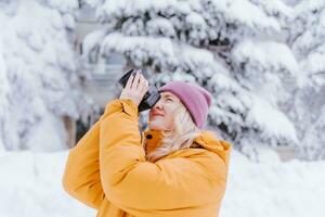 feliz menina fotógrafo dentro uma amarelo Jaqueta leva As fotos do inverno dentro uma Nevado parque