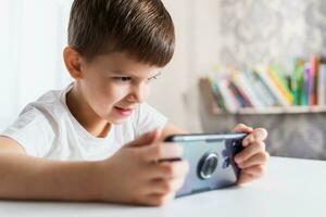 uma alegre criança dentro uma branco camiseta tocam jogos em a telefone às lar. feliz Garoto parece às dele Smartphone. foto