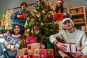 grupo do quatro jovem amigos vestido dentro vermelho chapéus e Natal camisolas dar presentes debaixo a Natal árvore foto