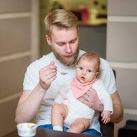 pai alimentando dele bebê fruta purê dentro a cozinha foto